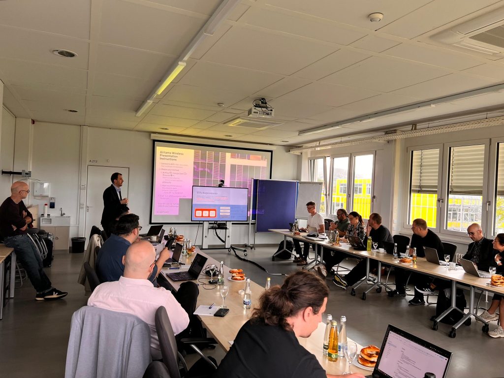 A group of WP2 representatives sit around a conference table at Hochschule Offenburg, working on laptops and listening to a presenter. A screen shows a slide about “WP2 Requirements.” Bottles of water and snacks are placed on the tables.