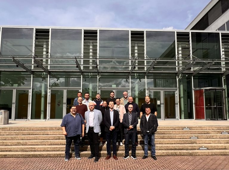 Group photo of WP2 representatives standing on the steps in front of a glass building at Hochschule Offenburg. About 15 people are posing together, smiling at the camera.