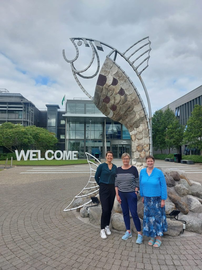 Three women stand in front of a large fish-shaped sculpture on the campus of ATU Sligo in Ireland. The word “WELCOME” is displayed in large white letters, and a modern glass building is visible in the background.