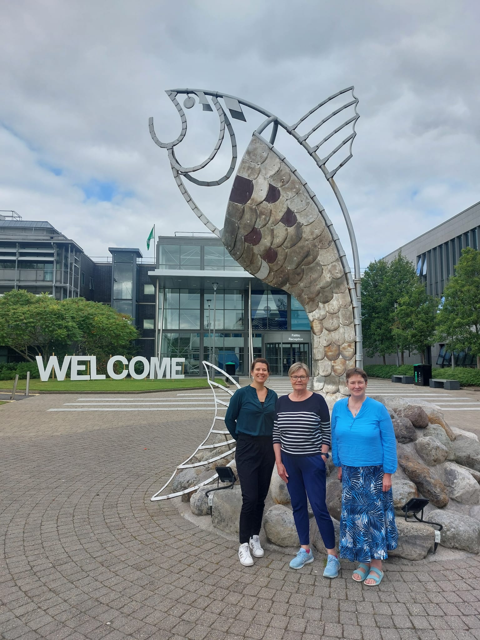 Three women stand in front of a large fish-shaped sculpture on the campus of ATU Sligo in Ireland. The word “WELCOME” is displayed in large white letters, and a modern glass building is visible in the background.