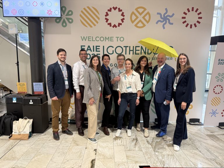 A group of ChallengeEU representatives posing together at EAIE Gothenburg 2025 in front of a welcome wall with colorful symbols. One person is holding a yellow umbrella, adding a cheerful touch to the group photo.