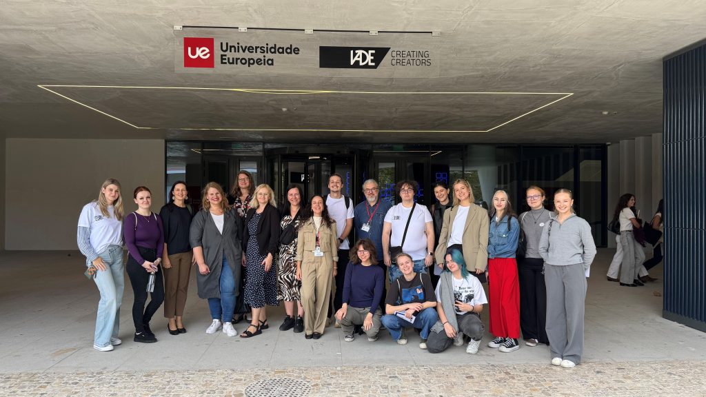 Group photo of students and staff posing in front of the Universidade Europeia / IADE – Creating Creators building entrance; the group stands under the campus sign, smiling toward the camera.