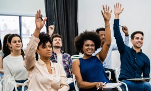 Students in a classroom raising their hands, smiling and engaged, some holding notebooks and pens