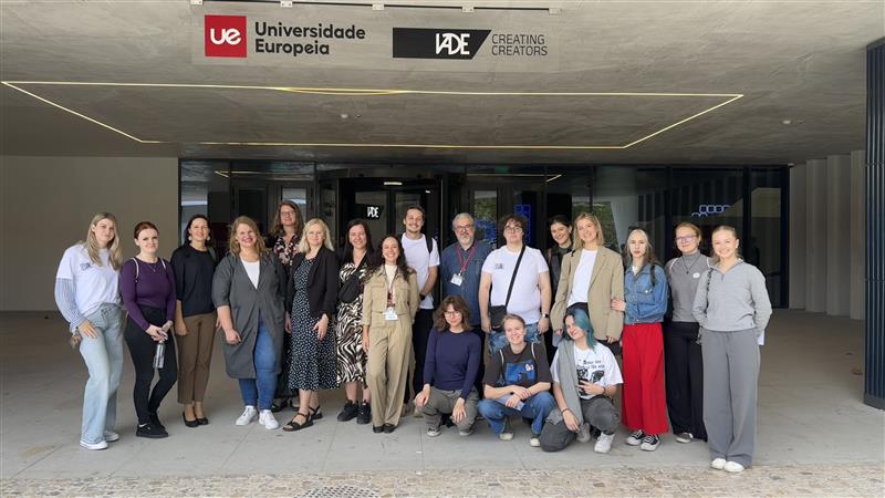 Group photo of students and staff posing in front of the Universidade Europeia / IADE – Creating Creators building entrance; the group stands under the campus sign, smiling toward the camera.