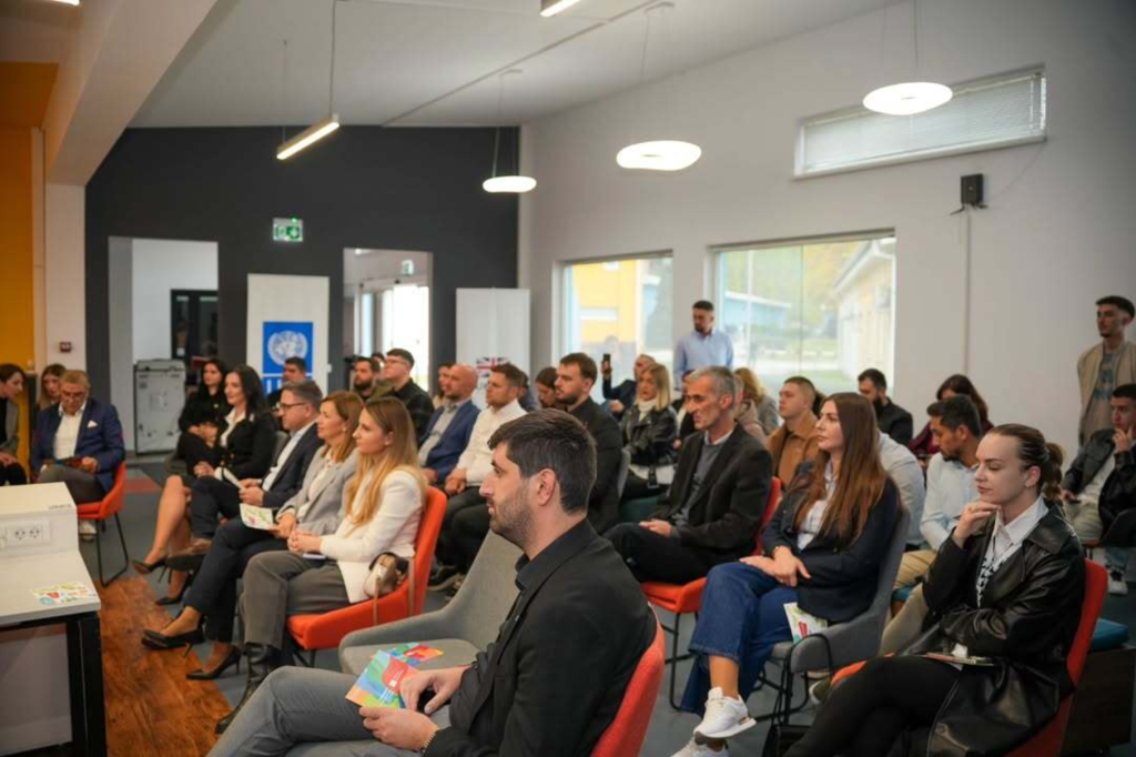 Attendees seated in a bright room listen attentively during the forum; event banners by the door and daylight from large windows.