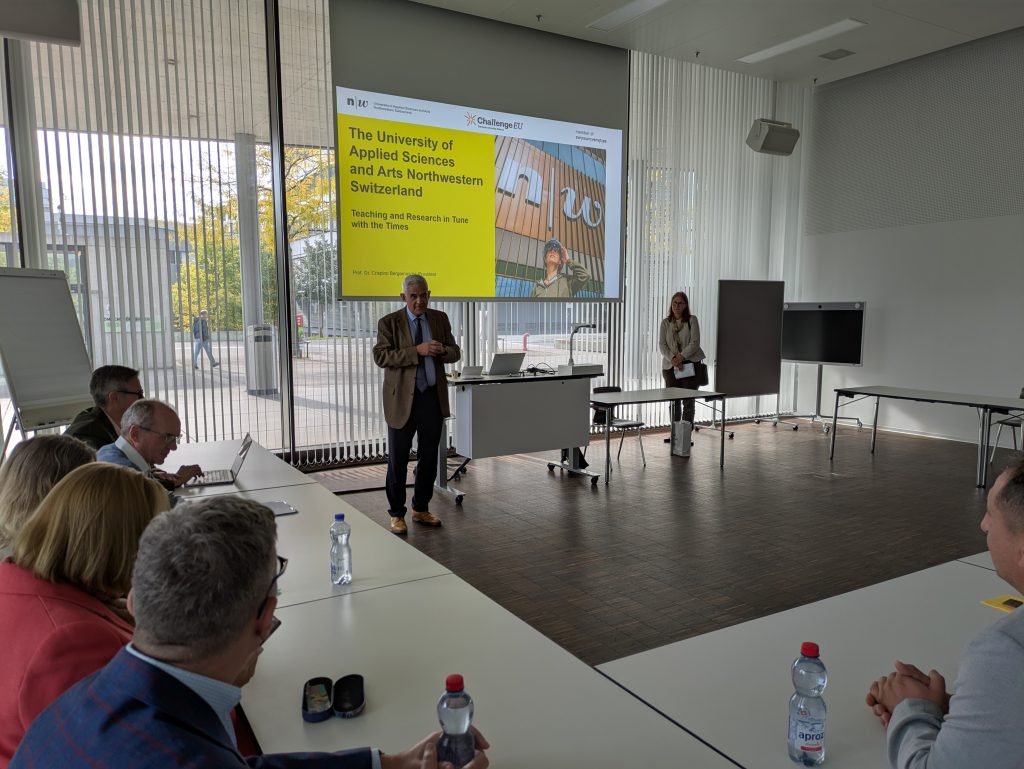 Wide view of a meeting room at FHNW with participants listening to a presenter; desks arranged in clusters