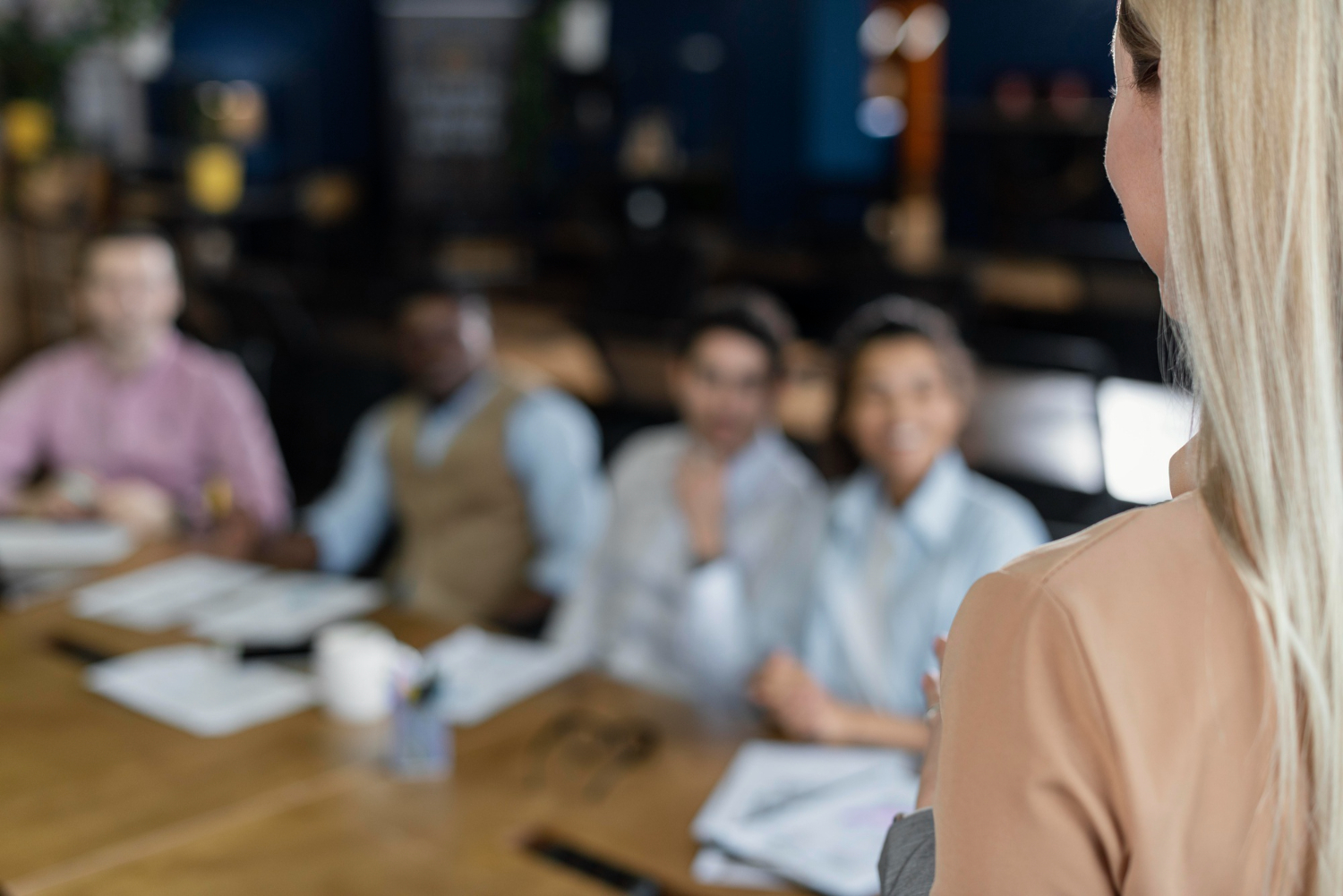 A speaker addresses a small group at a conference table; attendees sit blurred in the background, listening during a meeting/workshop.