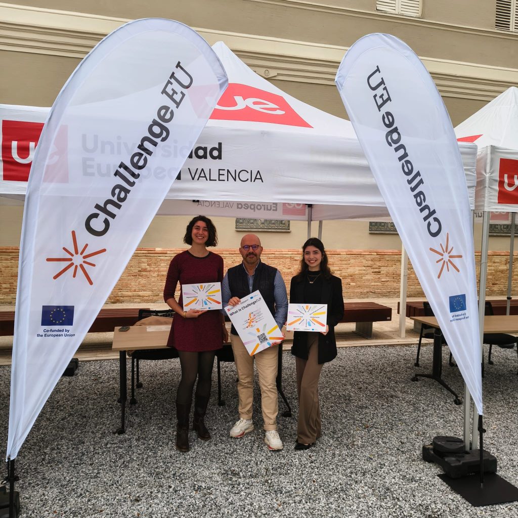 Three representatives stand under a Universidad Europea de Valencia tent between two ChallengeEU flags, holding ChallengeEU promotional materials in an outdoor courtyard, with the EU co-funding logo visible on the banners.