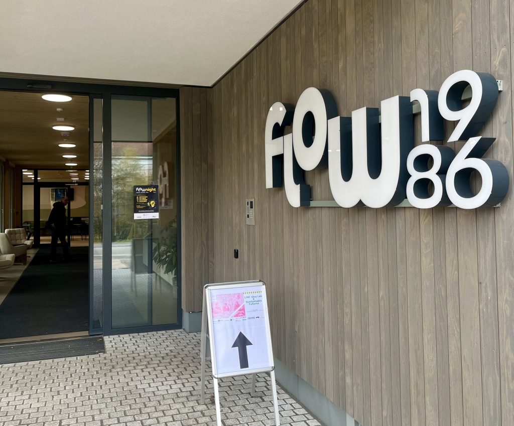 Entrance to the Flow 1986 coworking building with a large “flow1986” sign and a poster and arrow sign guiding participants to the Sustainable Idealab workshop