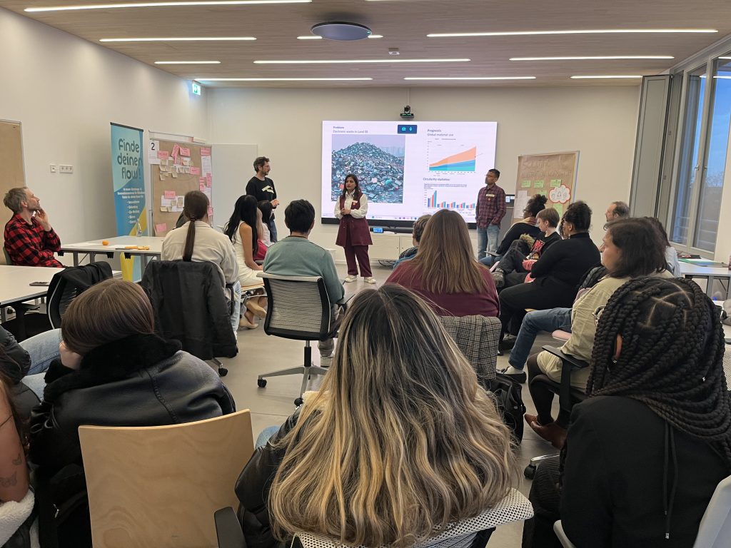 Participants seated in a workshop room listening to a team presenting their challenge solution in front of a large screen with graphs and images.