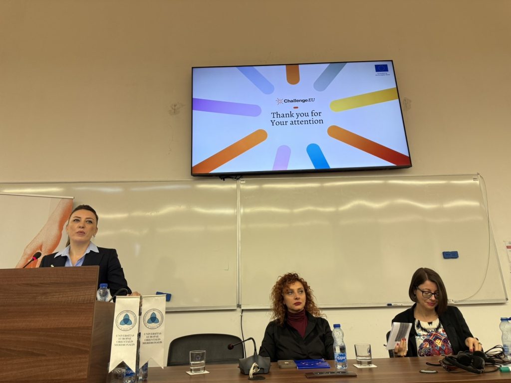 Speaker at the podium presenting during the ChallengeEU “Orange for Awareness” seminar, with two panelists seated at the table and the event slide displayed on the screen.