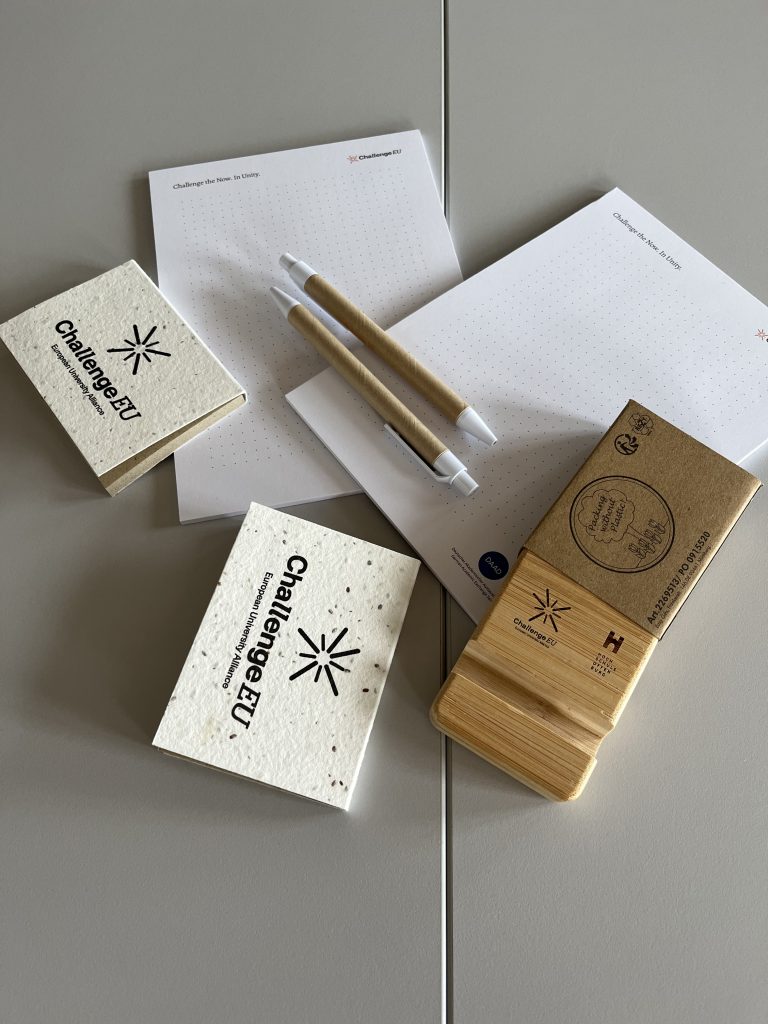 ChallengeEU-branded sustainable goodies on a table, including seed-paper notebooks, eco notepads, wooden phone stands and pens.