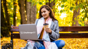 Young woman sitting on a park bench in an autumn setting, holding a takeaway coffee and working on a laptop with the ChallengeEU logo on the lid.