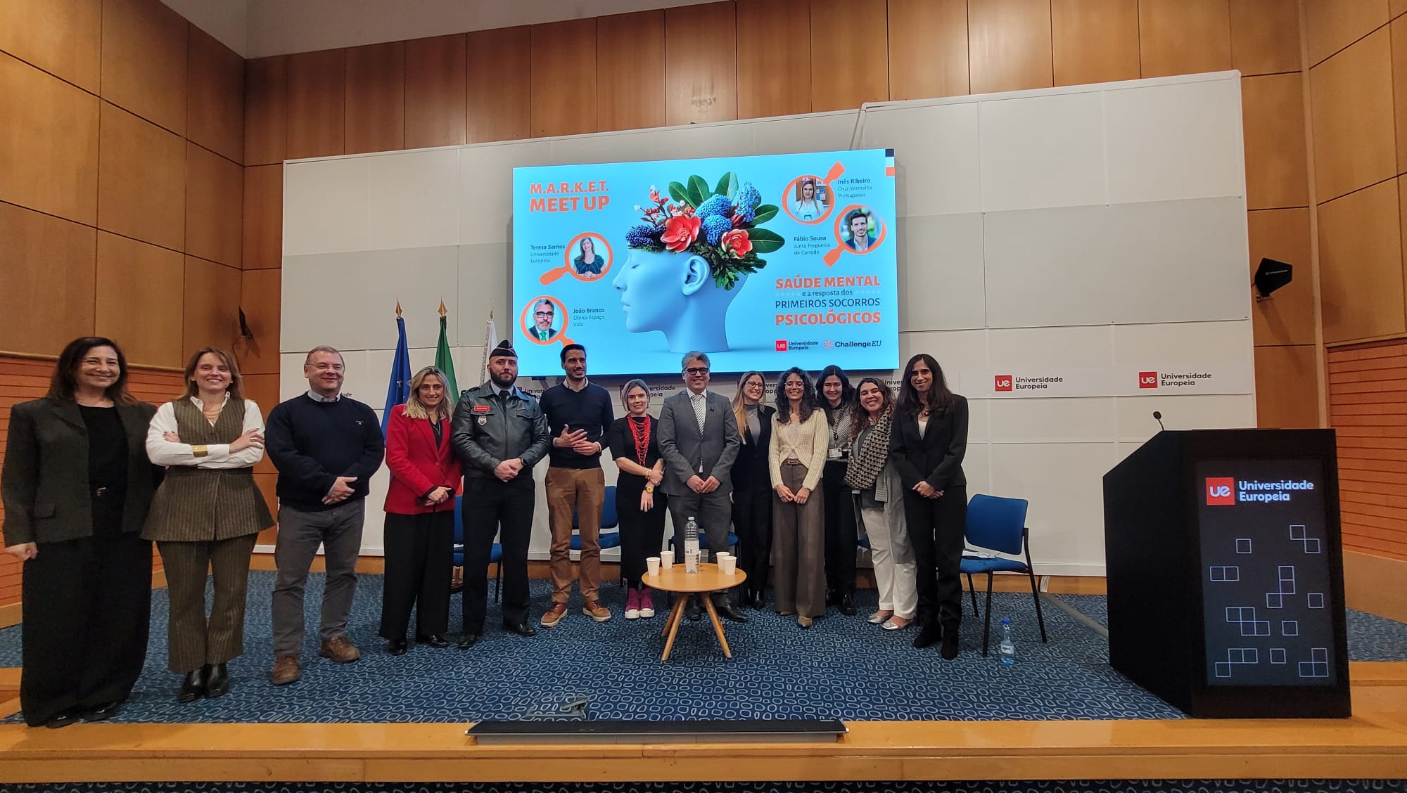 Group photo of participants on the auditorium stage beneath the M.A.R.K.E.T. Meet-Up slide on mental health and psychological first aid, with Universidade Europeia branding.