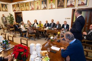 Wide view of the meeting room: a speaker addresses participants seated at U-shaped tables beneath portraits on the wall, with Christmas decorations and meeting materials visible.