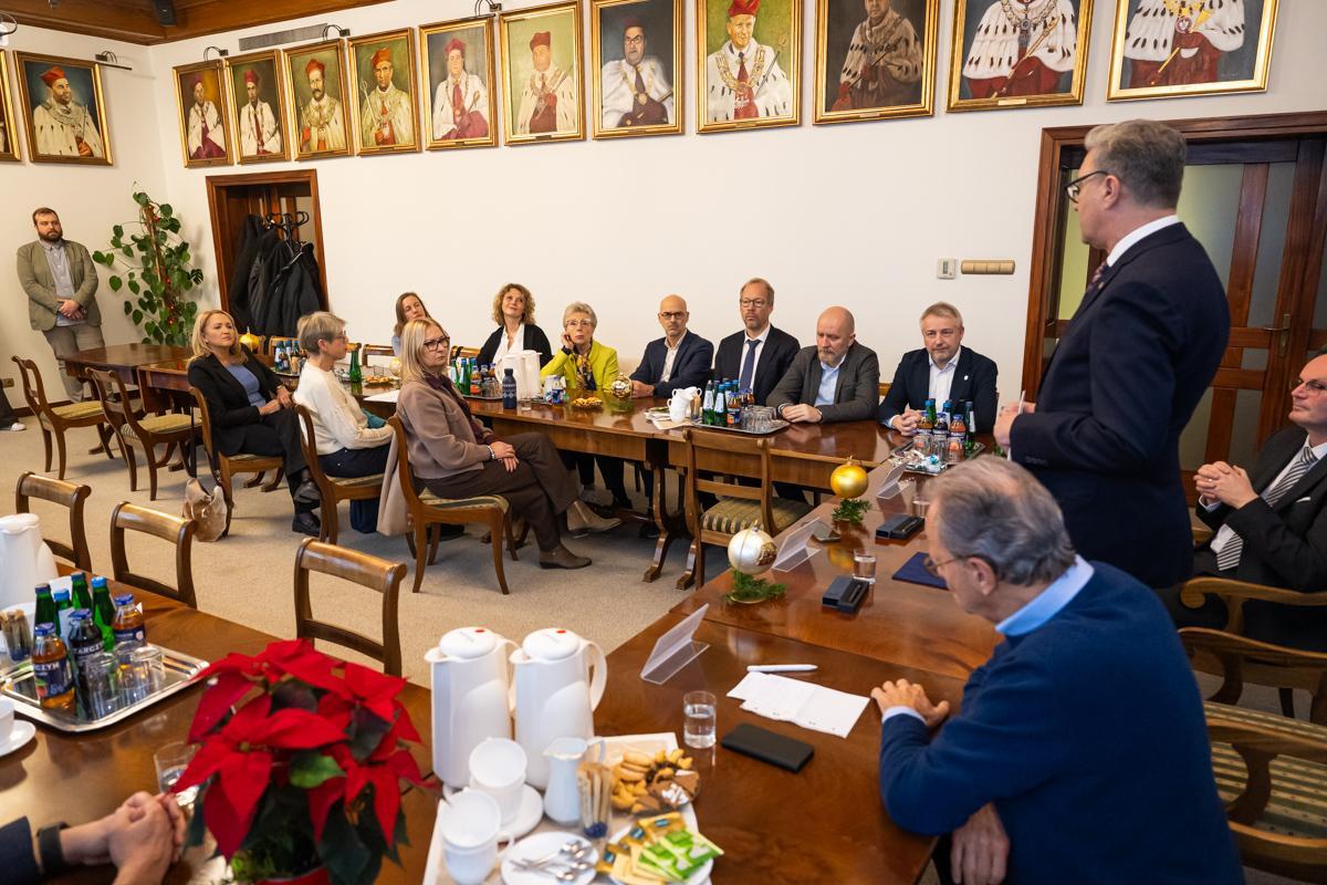Wide view of the meeting room: a speaker addresses participants seated at U-shaped tables beneath portraits on the wall, with Christmas decorations and meeting materials visible.