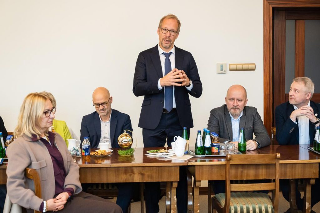 A speaker stands and addresses the group during the meeting; attendees sit at the table with notes, water bottles and festive décor.