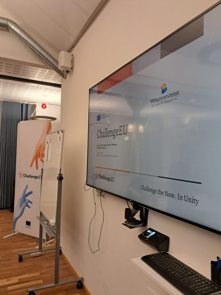 Close-up of a screen displaying a ChallengeEU presentation with Mid Sweden University (MIUN) branding; a whiteboard and roll-up banner stand nearby.