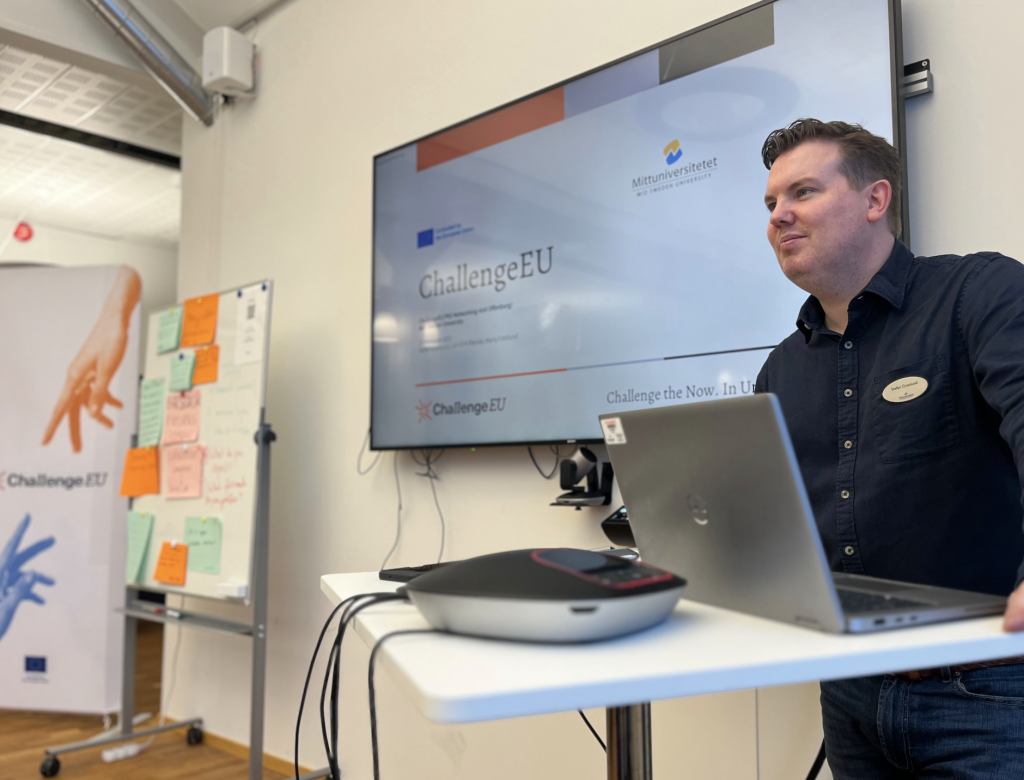 A speaker presents at a standing table with a laptop; behind him, a screen shows the ChallengeEU slide with MIUN branding and a whiteboard with sticky notes.