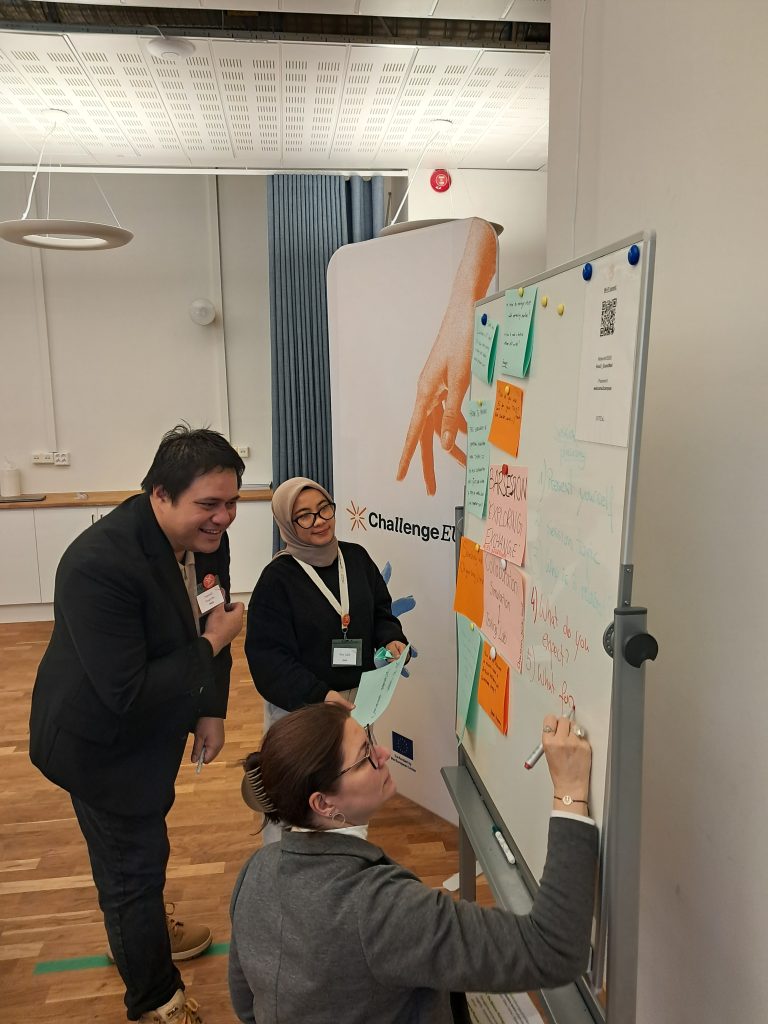 Three participants collaborate at a whiteboard covered with sticky notes; one person writes while others review ideas, with ChallengeEU branding behind.