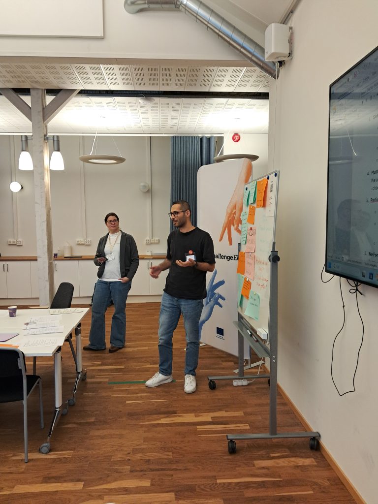 : A participant speaks during a session next to a whiteboard full of sticky notes, while another participant observes; workshop materials and a screen are visible.