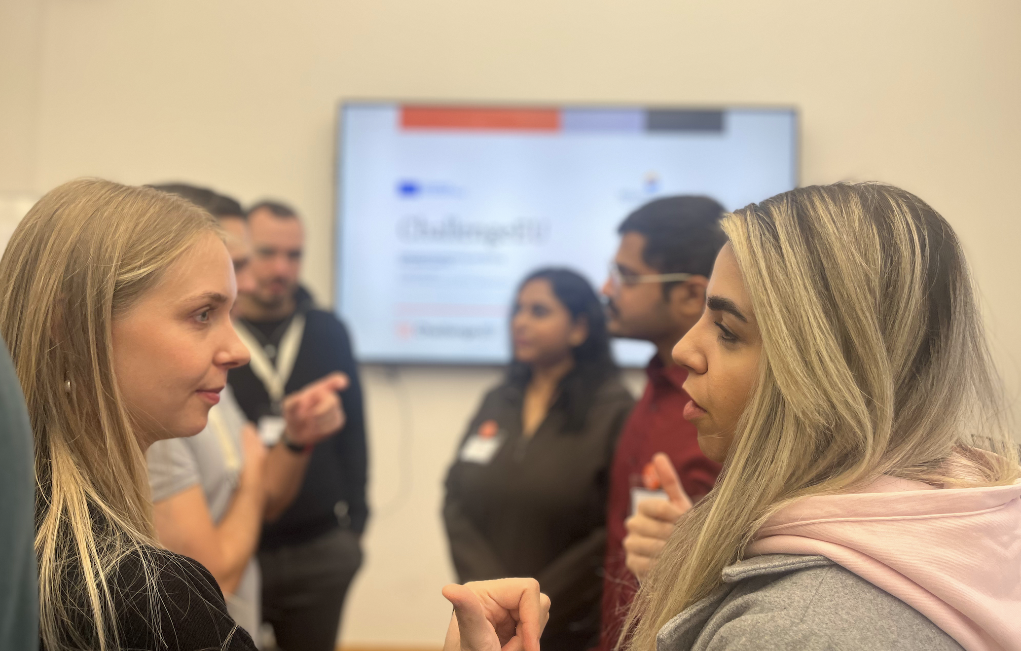 Two participants in conversation during the networking visit, with other attendees and a ChallengeEU slide blurred in the background.