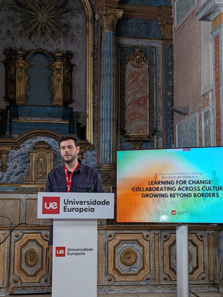 Speaker delivering a presentation at a podium in a historic university hall, with a presentation screen visible beside them.