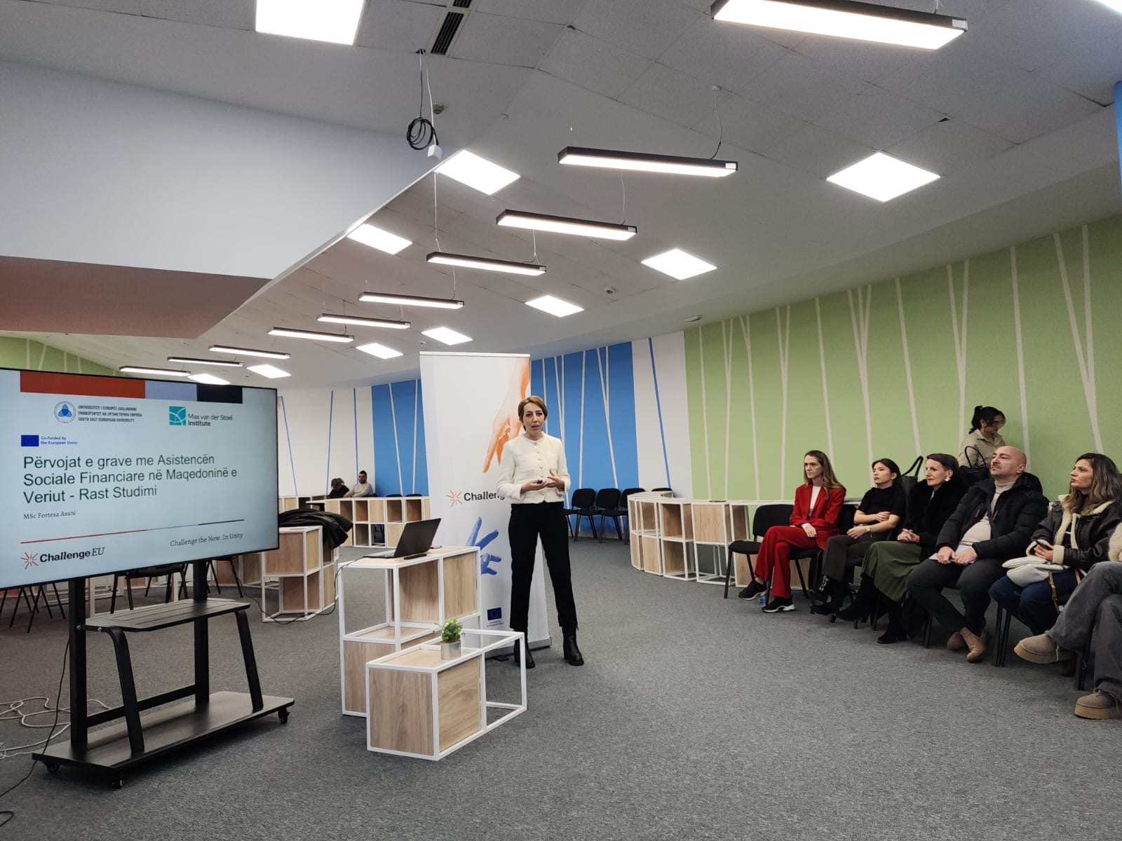 A presenter from SEEU stands next to a screen displaying a case study on the experiences of women using social assistance in North Macedonia; in front of her sits a group of participants attending the ChallengeEU seminar.