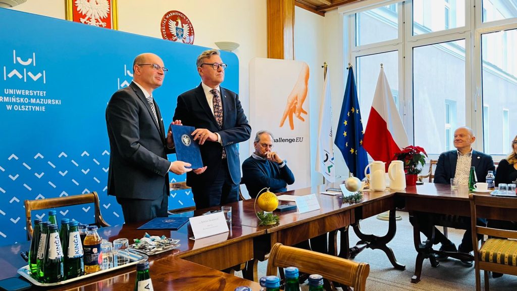 Two representatives pose in the UWM rectorate meeting room holding a blue document folder, with UWM and ChallengeEU banners and the EU and Polish flags in the background.