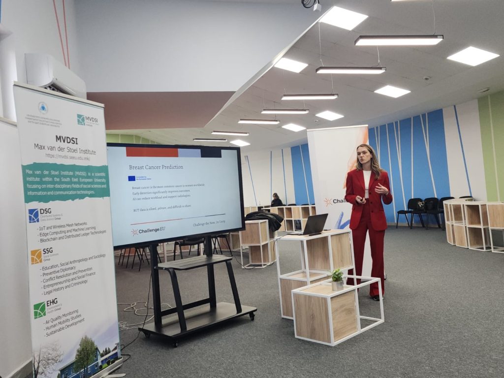 A presenter in a red suit delivers a presentation on breast cancer prediction; next to her is a Max van der Stoel Institute roll-up and the workshop room background.