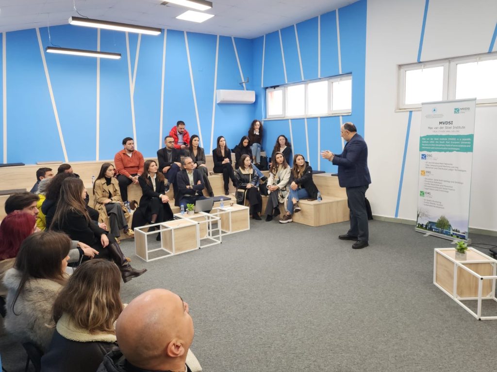 A group of students and academic staff sit on raised benches during a presentation led by a lecturer; an MDSI roll-up is visible in the room.