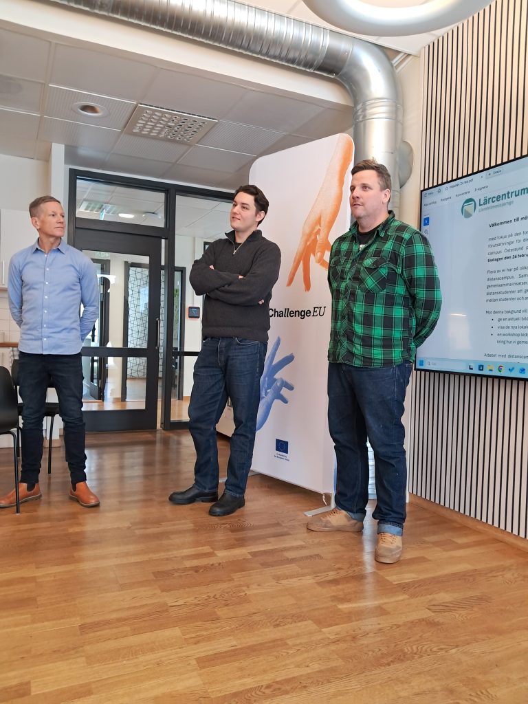 Three men stand beside a ChallengeEU roll-up banner and a large display screen showing a workshop agenda in Swedish. The men face an audience, listening attentively during the session.