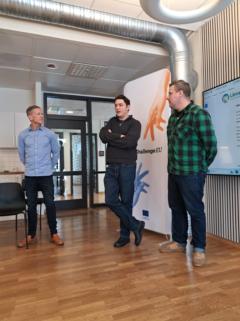 Three men stand beside a ChallengeEU roll-up banner and a large display screen showing a workshop agenda in Swedish. The men face an audience, listening attentively during the session.