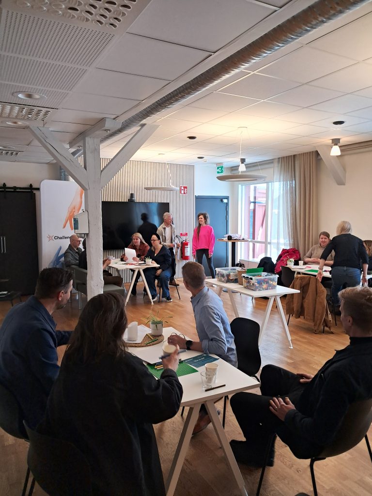 Participants sit and stand around several white tables during a ChallengeEU workshop session. LEGO bricks, green baseplates and coffee cups are placed on the tables. A roll-up banner and a large screen are visible in the background as small groups discuss their work.