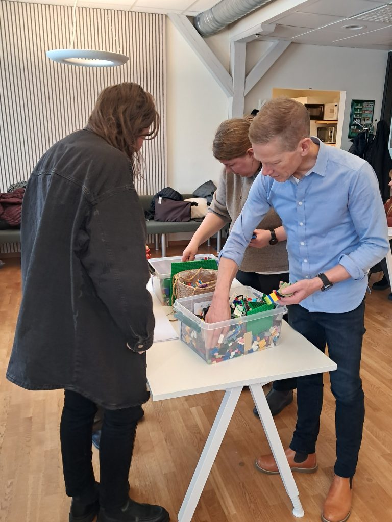 Group participants collaborate at a table, building a small LEGO model on a green baseplate. Coloured markers, notes and small figurines are spread across the table. A ChallengeEU banner stands next to a large black screen in the background.