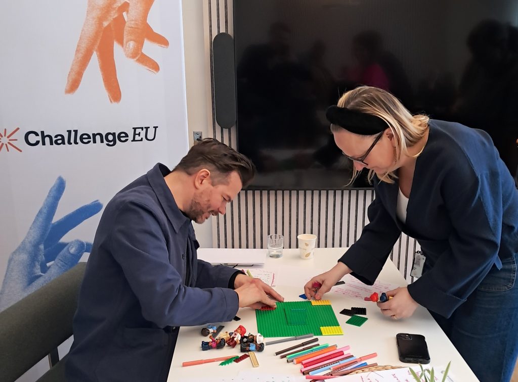 Two participants collaborate at a table, building a small LEGO model on a green baseplate. Coloured markers, notes and small figurines are spread across the table. A ChallengeEU banner stands next to a large black screen in the background.