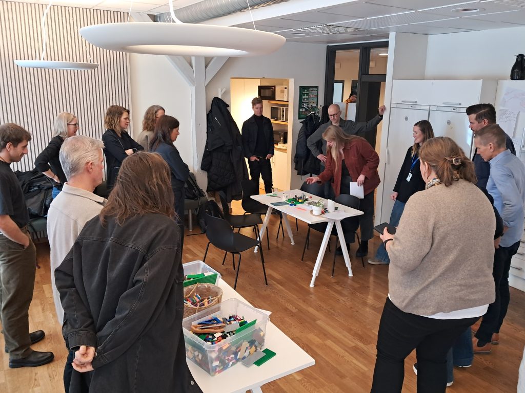 Approximately a dozen participants form a circle around tables with LEGO bricks during a workshop session. One woman leans forward to place a piece on a LEGO model while others observe. The room features wooden flooring and white walls.