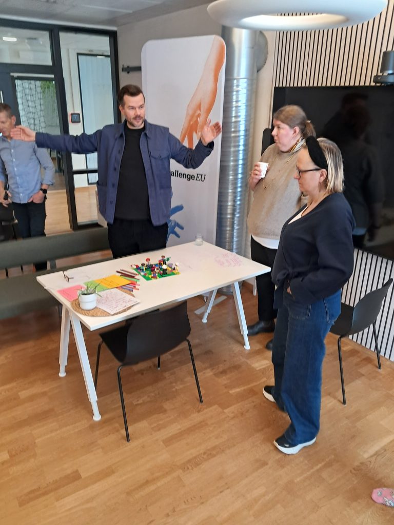 A facilitator stands with arms extended while explaining an activity to three participants gathered around a white table with LEGO bricks, markers and notes. A ChallengeEU roll-up banner and a large screen are visible behind them.