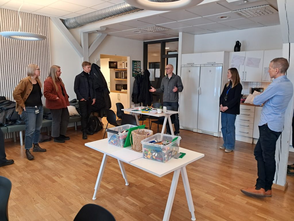 A group of adults standing in a bright workshop room arranged around two white tables with containers of LEGO bricks. A man in glasses gestures while speaking. Coats hang on a rack behind them, and a small kitchenette is visible in the background.