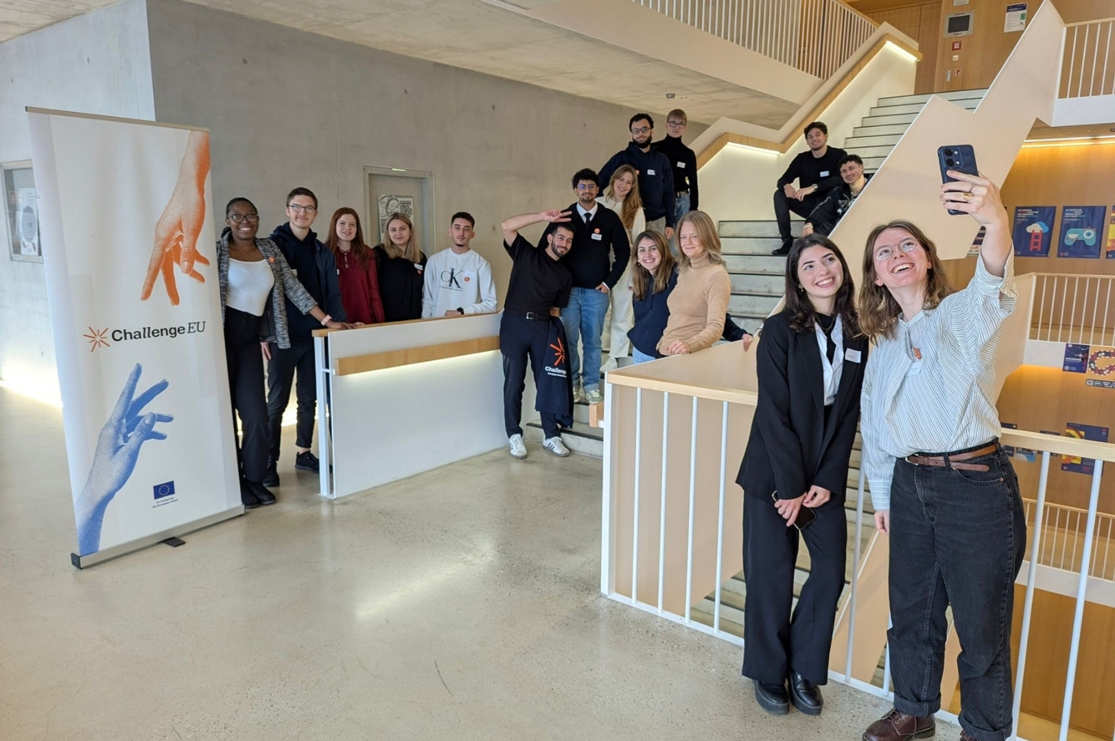 A group of international students stands together on a staircase inside a modern university building. They are smiling and posing for a group photo, with two students in the foreground taking a selfie. A Chal