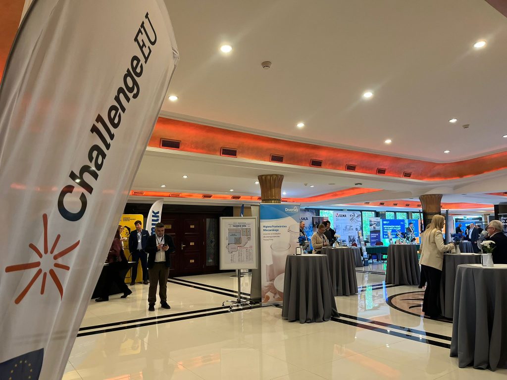 Close-up of ChallengeEU flag inside the Techmilk venue, with conference space and attendees in the background.