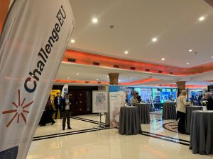 Close-up of ChallengeEU flag inside the Techmilk venue, with conference space and attendees in the background.