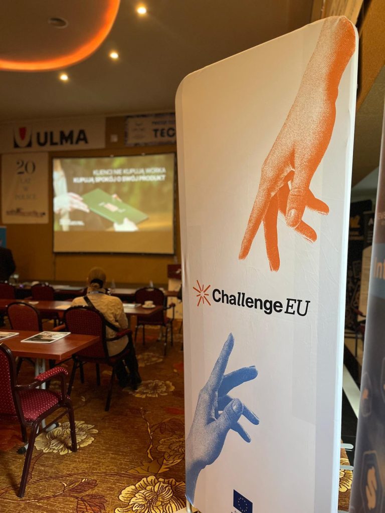ChallengeEU roll-up banner displayed inside the Techmilk conference hall, with presentation screen and seating area in the background.