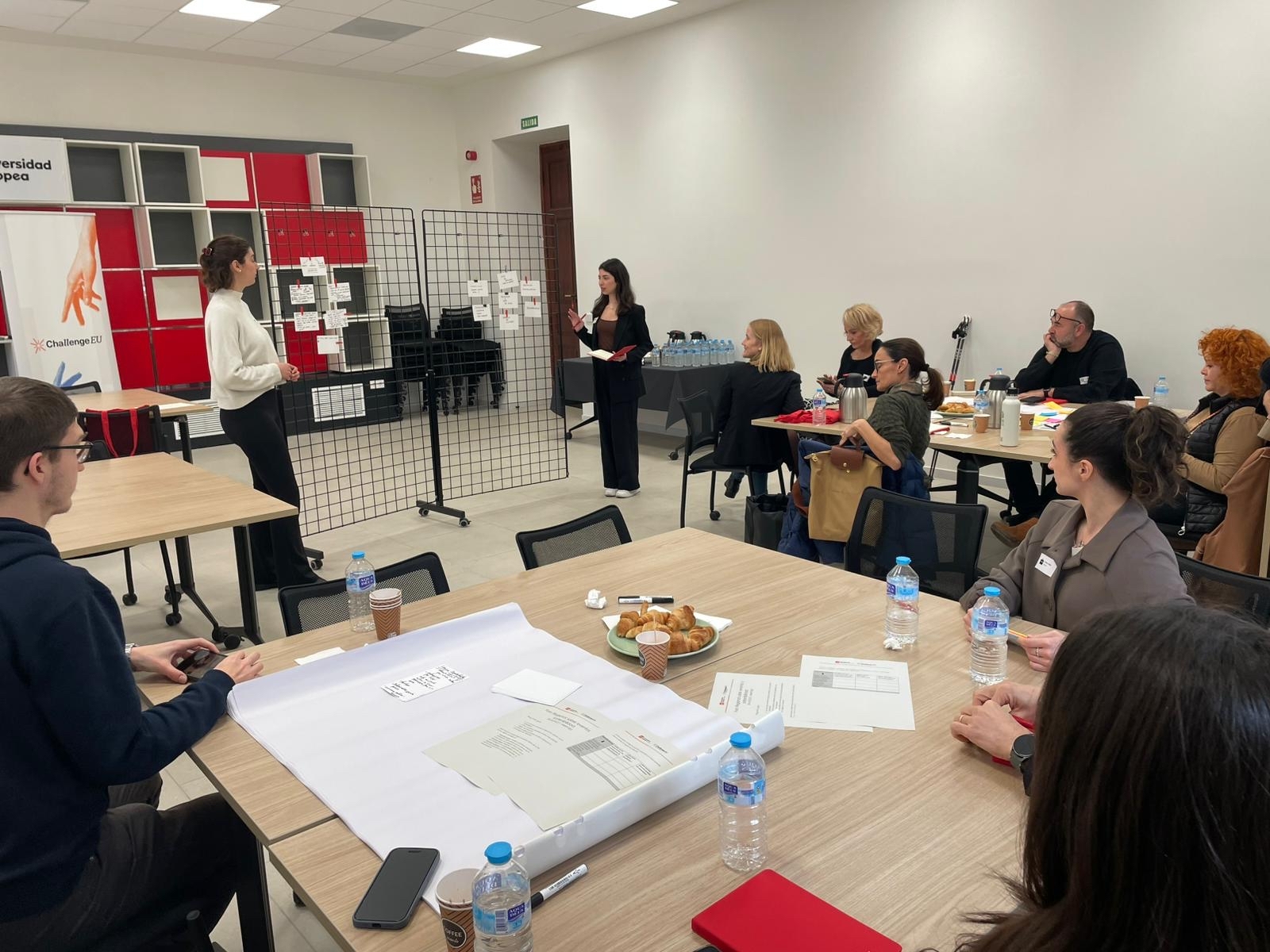 Participants attend the M.A.R.K.E.T. Meet-Up workshop on Events and Sustainability, engaging in group discussions around tables while two facilitators present ideas at the front of the room.