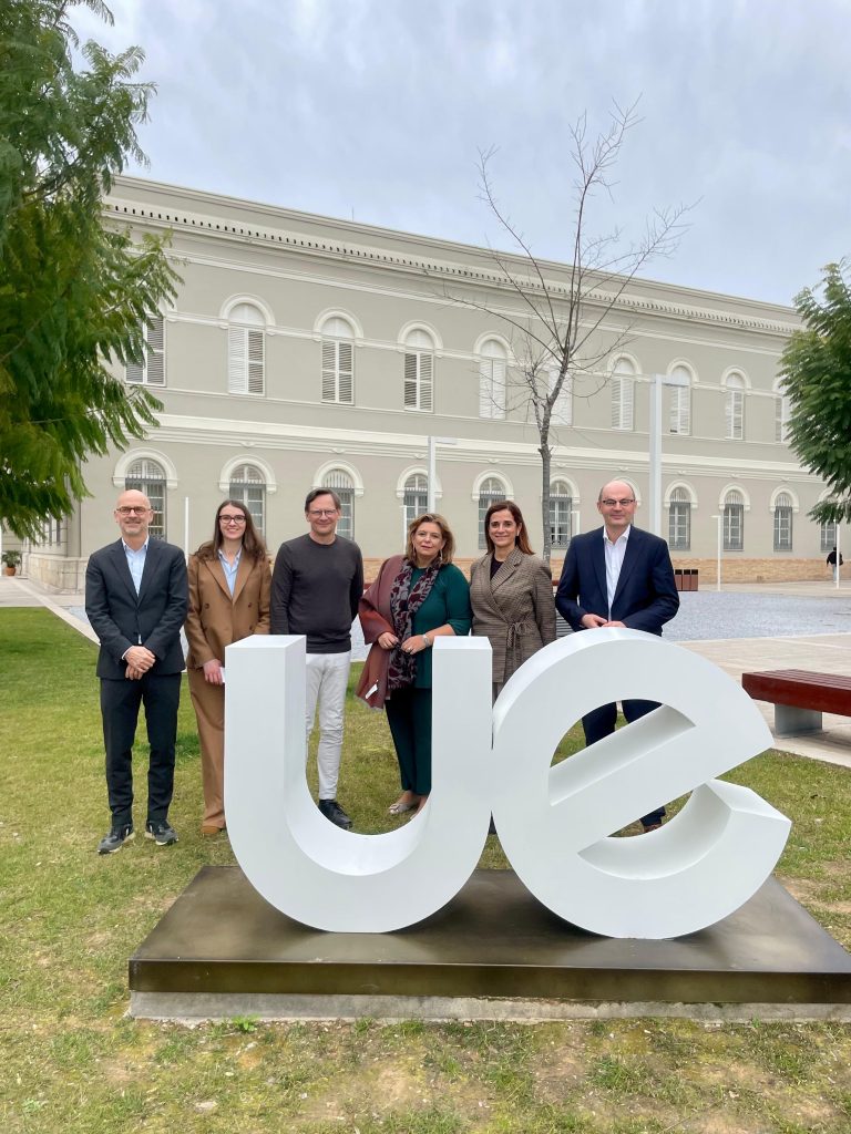 Group photo with the host Rector from Valencia together with the Rectors from Hochschule Offenburg (HSO), Universidade Europeia Lisbon (UEL) and Mid Sweden University (MIUN), standing outdoors in front of the university building and large “UE” letters.
