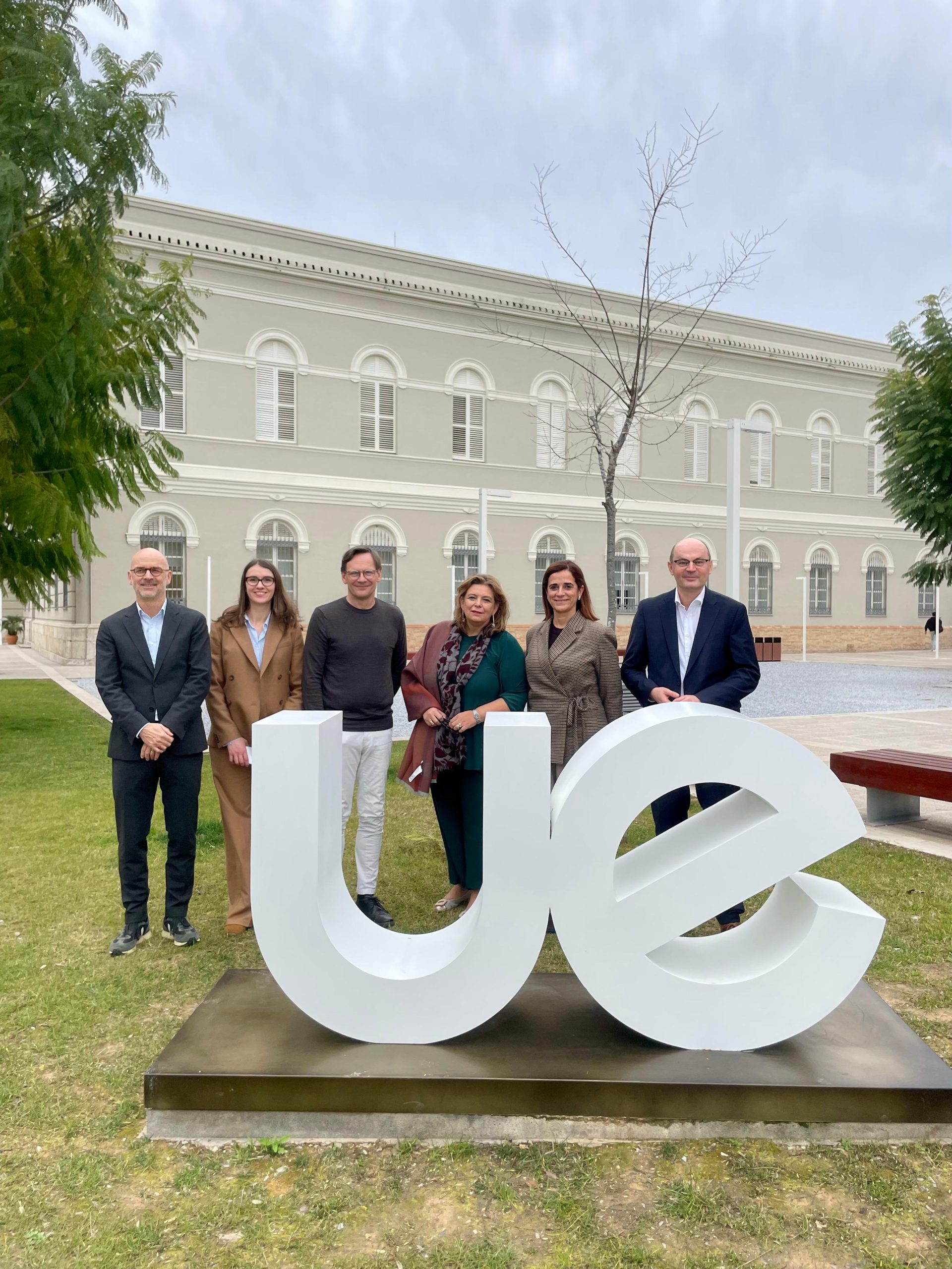Group photo with the host Rector from Valencia together with the Rectors from Hochschule Offenburg (HSO), Universidade Europeia Lisbon (UEL) and Mid Sweden University (MIUN), standing outdoors in front of the university building and large “UE” letters.