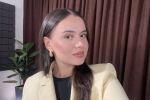 Portrait of a young woman with long dark hair, wearing a light beige blazer, black top, and gold earrings, posing indoors in front of dark curtains with a plant in the background.