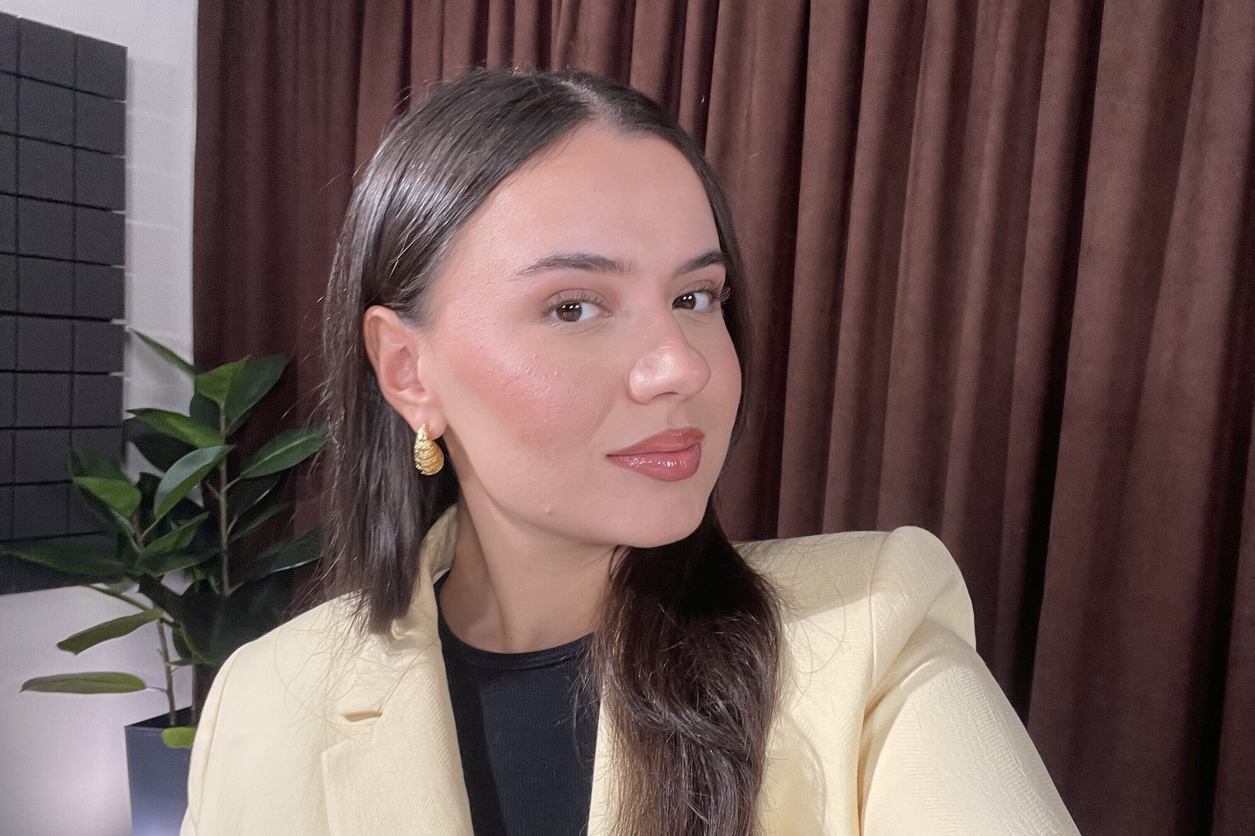 Portrait of a young woman with long dark hair, wearing a light beige blazer, black top, and gold earrings, posing indoors in front of dark curtains with a plant in the background.