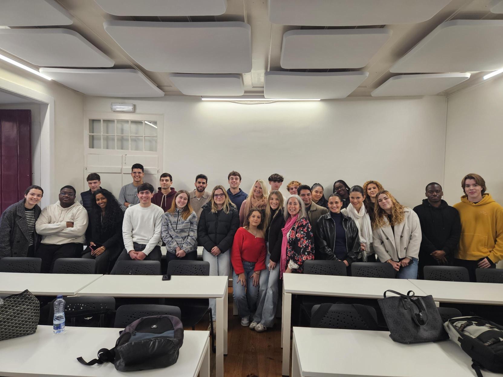 A group of international university students and staff posing together in a classroom with desks and acoustic ceiling panels, following a meeting or academic session.