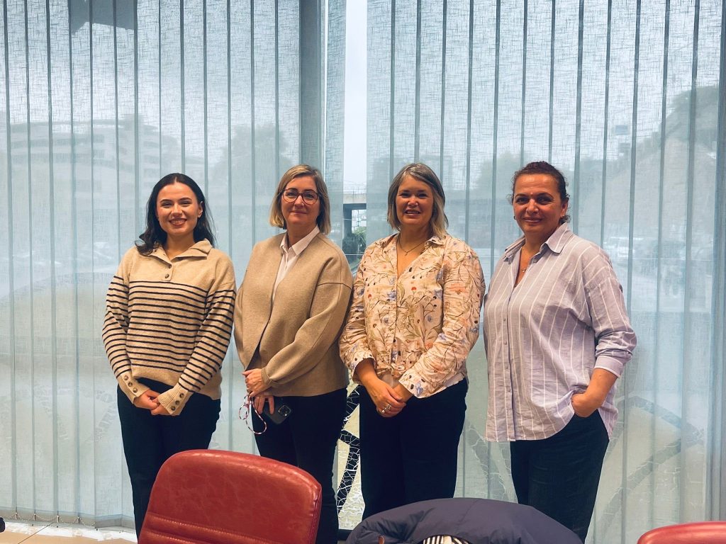Four women standing side by side in a meeting room in front of large windows with vertical blinds, posing for a group photo.